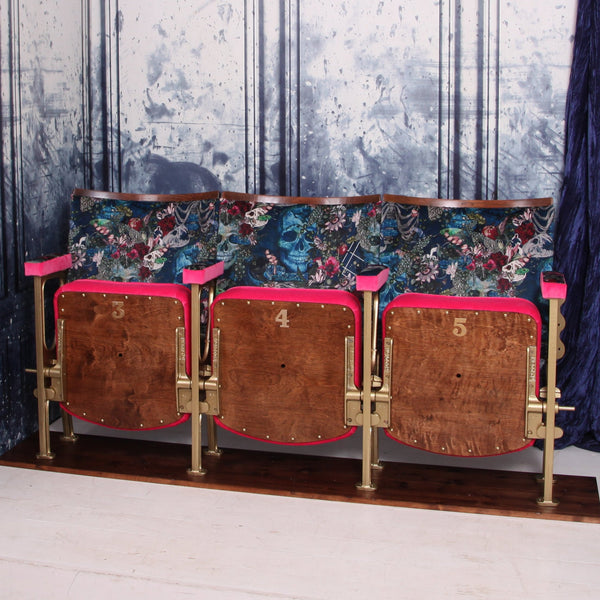 Set of three vintage cinema seats with seats up upholstered in blue skull velvet with pink piping and panels.  The seats are in front of a blue marbled wall.