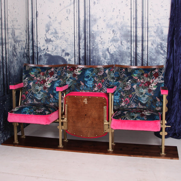 Set of three vintage cinema seats with two seats down and one up, upholstered in blue skull velvet with pink piping and panels.  The seats are in front of a blue marbled wall.