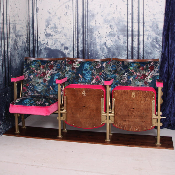 Set of three vintage cinema seats with one seat down, upholstered in blue skull velvet with pink piping and panels.  The seats are in front of a blue marbled wall.