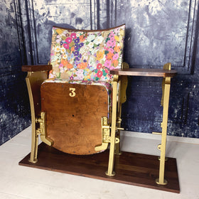 a single vintage cinema seat upholstered a multicoloured floral linen with a side table attached.  Seat is sat in front of a blue marbled wall.11