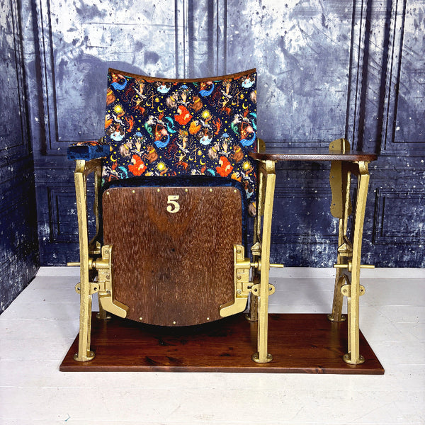 Vintage cinema seat with seat closed, upholstered in a dark blue velvet featuring zodiac themed velvet , against a blue marbled wall.