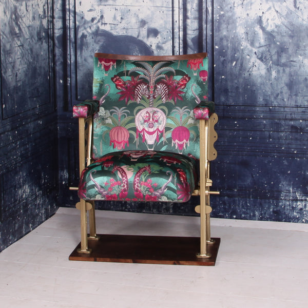 A SINGLE VINTAGE CINEMA SEAT UPHOLSTERED IN A PINK AND GREEN VELVET WITH ANIMALS AND AIR BALLOONS ON IT.  THE CHAIR IS IN FRONT OF A BLUE WALL