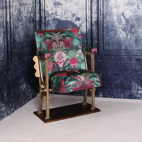 A SINGLE VINTAGE CINEMA SEAT UPHOLSTERED IN A PINK AND GREEN VELVET WITH ANIMALS AND AIR BALLOONS ON IT.  THE CHAIR IS IN FRONT OF A BLUE WALL