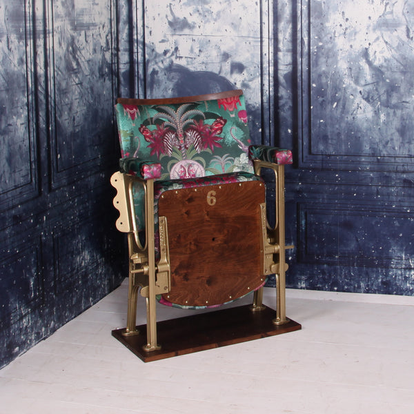 A SINGLE VINTAGE CINEMA SEAT UPHOLSTERED IN A PINK AND GREEN VELVET WITH ANIMALS AND AIR BALLOONS ON IT.  THE CHAIR IS IN FRONT OF A BLUE WALL