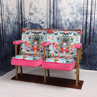 Set of two vintage cinema seats against a blue marbled wallpaper.  Seats are upholstered in a flower and butterfly pattern blue velvet with pink piping and panelling