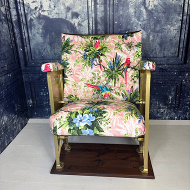A vintage cinema seat refurbished and upholstered in a pink velvet with scarlet parrots on it.  Shown on a white floor against a blue marbled background.