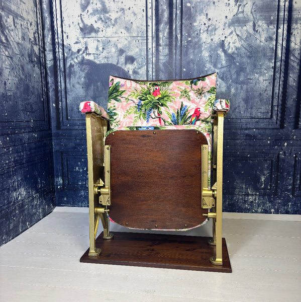A vintage cinema seat refurbished and upholstered in a pink velvet with scarlet parrots on it.  Shown on a white floor against a blue marbled background.