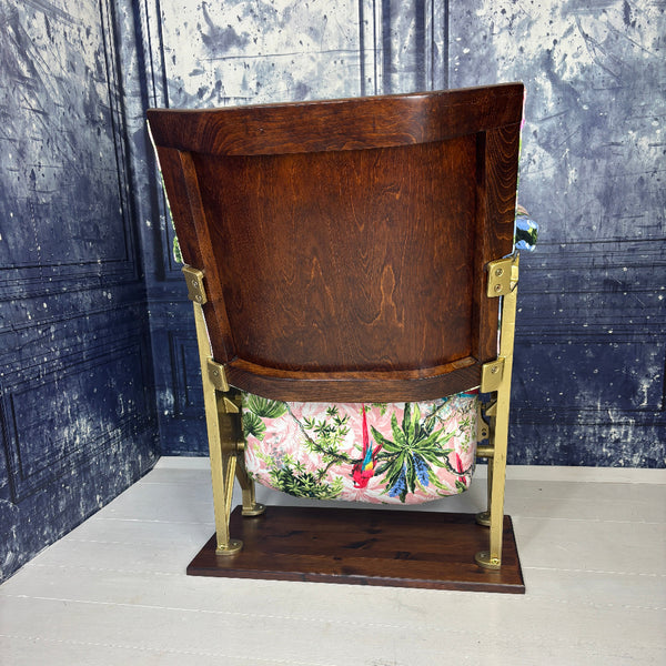 Rear view of a vintage cinema seat refurbished and upholstered in a pink velvet with scarlet parrots on it.  Shown on a white floor against a blue marbled background.
