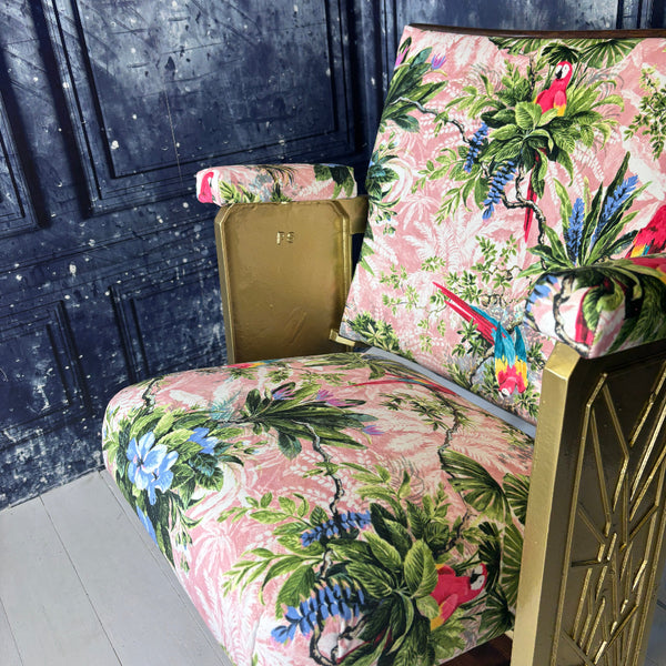 Close up of a vintage cinema seat refurbished and upholstered in a pink velvet with scarlet parrots on it.  Shown on a white floor against a blue marbled background.